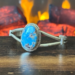 May include: A silver cuff bracelet featuring a large, oval-shaped blue turquoise stone with brown matrix inclusions. The stone is set in a serrated silver bezel, and the bracelet band consists of two smooth silver wires that meet at the sides.