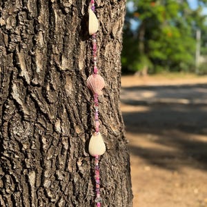 May include: A pink and white seashell garland with pink and clear beads hanging from a tree branch.