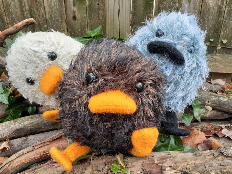 Peut inclure: Trois jouets en peluche en forme d'oiseaux. L'oiseau &agrave; gauche est blanc avec des yeux noirs et un bec orange. L'oiseau au centre est noir avec des plumes brunes, un bec orange et des yeux noirs. L'oiseau &agrave; droite est bleu avec des yeux noirs et un bec noir.