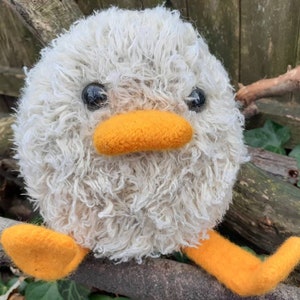Peut inclure: Un animal en peluche blanc et moelleux avec des pieds orange et un bec. L'animal a des yeux noirs et est assis sur une branche.