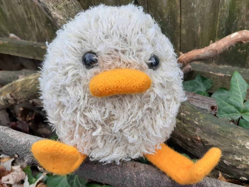 Peut inclure: Un animal en peluche blanc et moelleux avec des pieds orange et un bec. L'animal a des yeux noirs et est assis sur une branche.