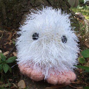 May include: A white fluffy crocheted toy with pink feet and black eyes. The toy is sitting on a brown surface with green plants in the background.