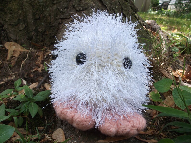 May include: A white fluffy crocheted toy with pink feet and black eyes. The toy is sitting on a brown surface with green plants in the background.