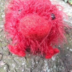 May include: A handmade, red, fuzzy creature with black bead eyes and small feet. The plush toy is made of a soft, textured material and is sitting on a tree trunk. The creature has a round body and a prominent, rounded nose.