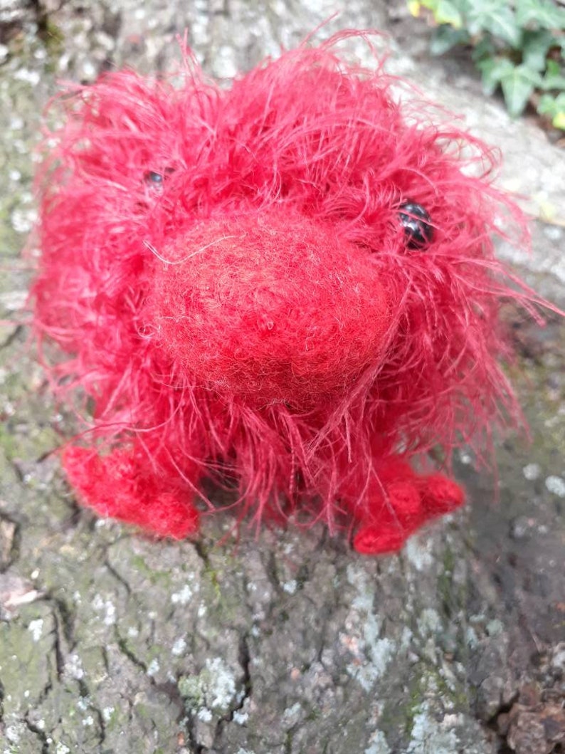 May include: A handmade, red, fuzzy creature with black bead eyes and small feet. The plush toy is made of a soft, textured material and is sitting on a tree trunk. The creature has a round body and a prominent, rounded nose.