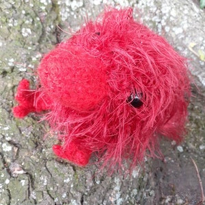 May include: A handmade, bright red, fuzzy creature with small, black eyes and tiny feet. The plush toy is made of a soft, textured material and is resting on a textured, gray and brown tree trunk.