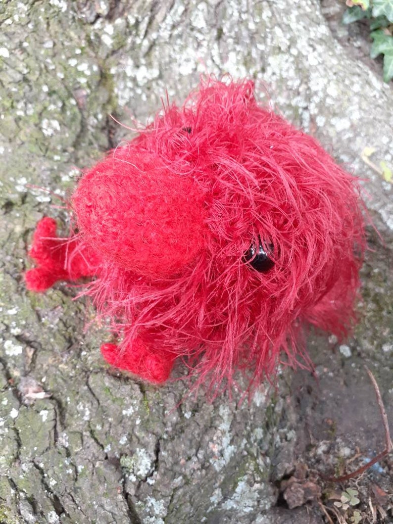 May include: A handmade, bright red, fuzzy creature with small, black eyes and tiny feet. The plush toy is made of a soft, textured material and is resting on a textured, gray and brown tree trunk.