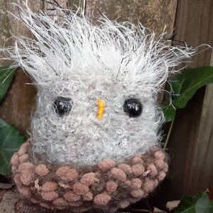 Pu&ograve; includere: Un gufo in crochet grigio e marrone con grandi occhi neri e un becco giallo. Il gufo &egrave; seduto su una superficie di legno con foglie verdi sullo sfondo.