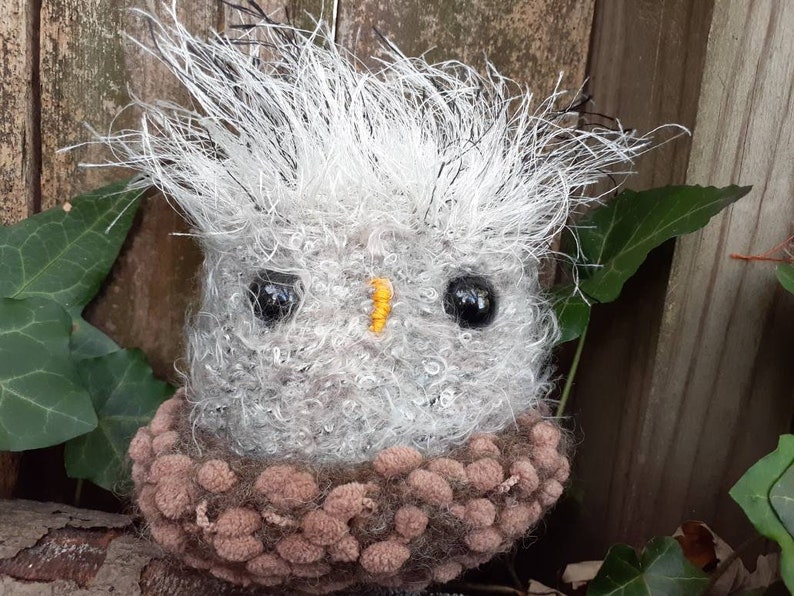 Pu&ograve; includere: Un gufo in crochet grigio e marrone con grandi occhi neri e un becco giallo. Il gufo &egrave; seduto su una superficie di legno con foglie verdi sullo sfondo.