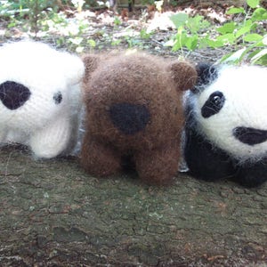 May include: Three knitted stuffed animals: a white polar bear, a brown bear, and a black and white panda bear. The animals are sitting on a wooden surface.