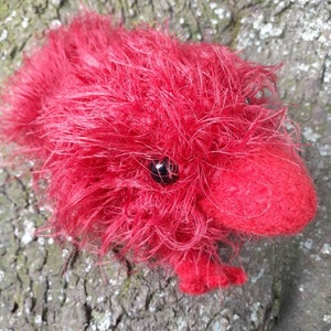 May include: A handmade red plush toy shaped like a small animal, possibly a hedgehog or a similar creature. The toy has a fluffy, textured appearance with a dark eye and small feet. The toy is resting on a textured, brown surface.