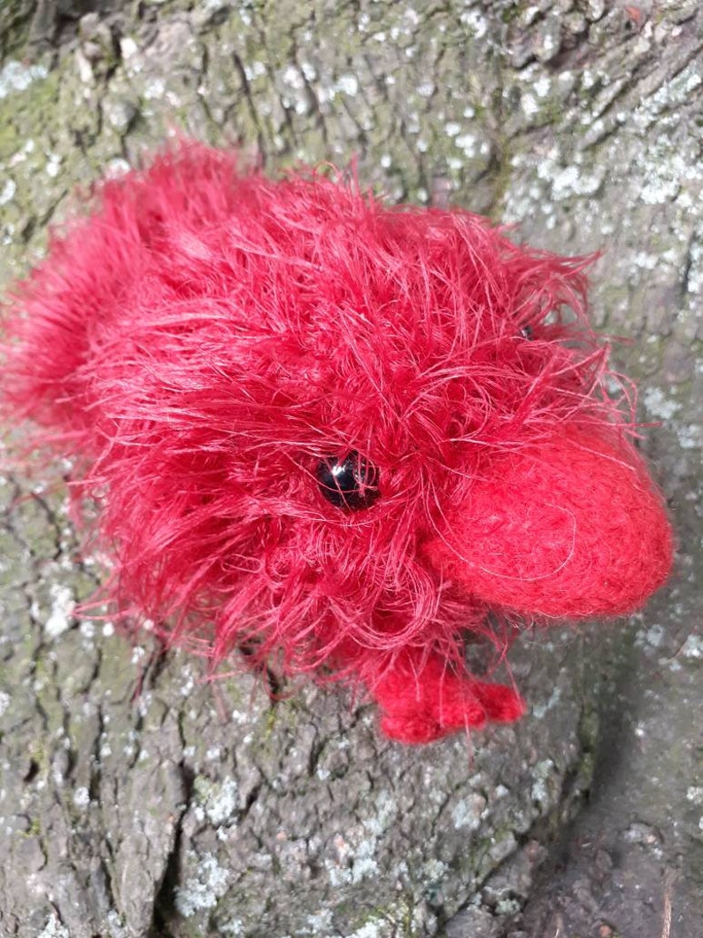 May include: A handmade red plush toy shaped like a small animal, possibly a hedgehog or a similar creature. The toy has a fluffy, textured appearance with a dark eye and small feet. The toy is resting on a textured, brown surface.