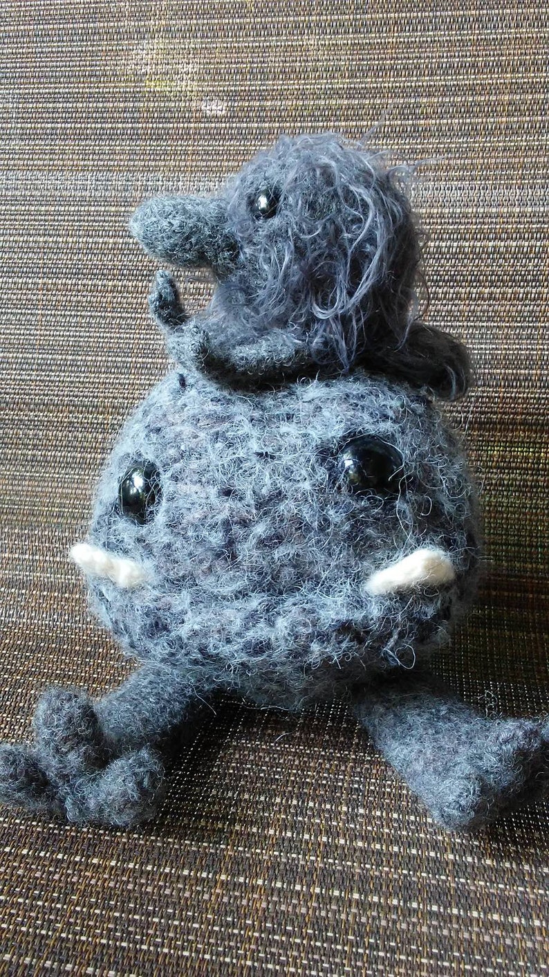 May include: A handmade gray plush creature with large black eyes, white tusks, and small arms and legs. A smaller creature sits on top of the larger one. The plush toy is made of a fuzzy material.