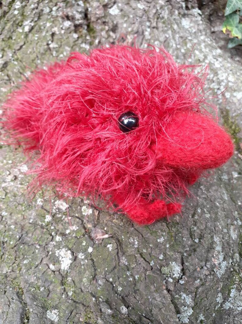 May include: A handmade, plush red creature with a fluffy, textured body and a small, black eye. The soft toy is lying on a textured, brown surface, possibly a tree trunk. The creature's design is whimsical and unique.