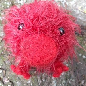 May include: A handmade, fuzzy red creature with black bead eyes, a rounded nose, and small feet. The plush toy is made of soft, textured material and is set against a natural, wood-like background. A unique, whimsical creation.