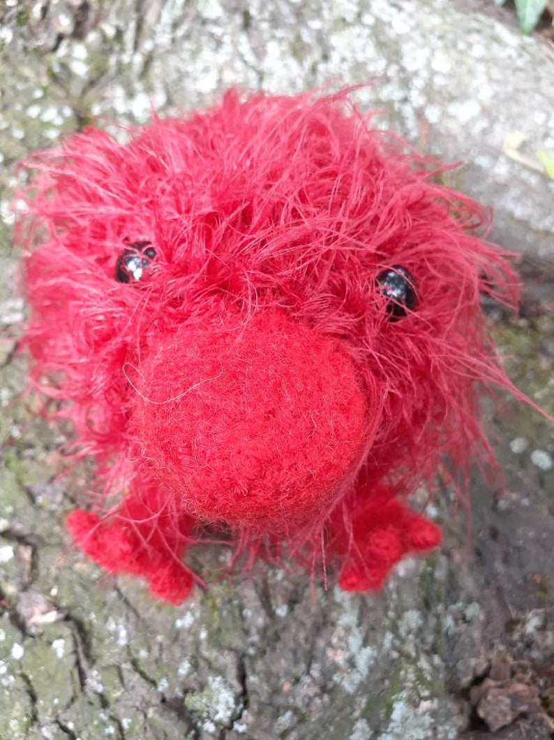 May include: A handmade, fuzzy red creature with black bead eyes, a rounded nose, and small feet. The plush toy is made of soft, textured material and is set against a natural, wood-like background. A unique, whimsical creation.