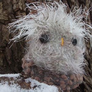 Pu&ograve; includere: Un gufo di peluche bianco e grigio a forma di gufo con occhi neri e un becco giallo. Il gufo &egrave; seduto su un tronco d'albero marrone con la neve a terra.