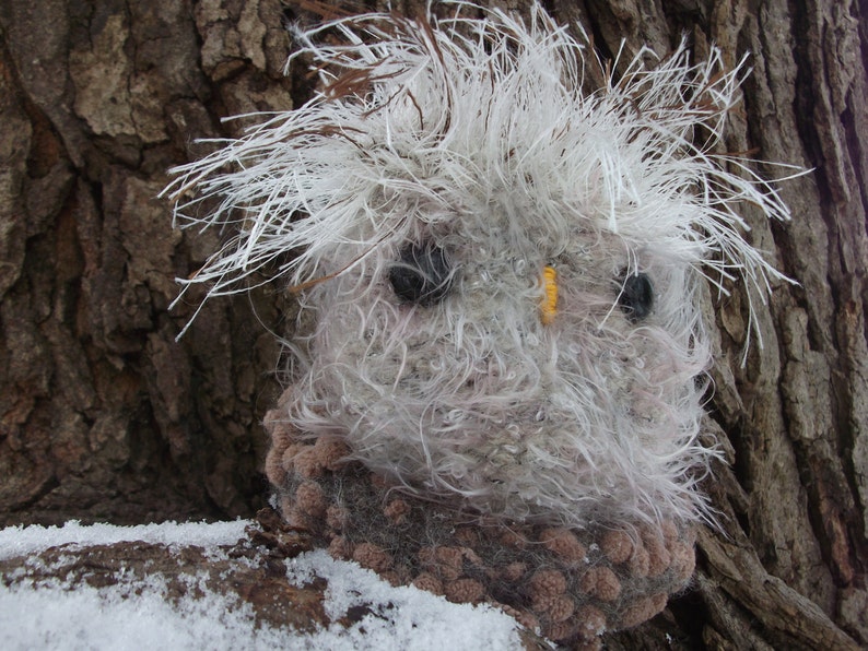 Pu&ograve; includere: Un gufo di peluche bianco e grigio a forma di gufo con occhi neri e un becco giallo. Il gufo &egrave; seduto su un tronco d'albero marrone con la neve a terra.