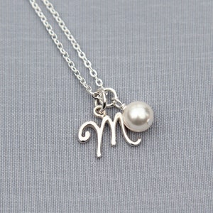 Puede incluir: Un collar de plata con un dije de letra "M" en cursiva y un colgante de perla blanca.