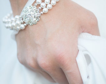 May include: A three-strand pearl bracelet with a decorative silver-tone centerpiece. The bracelet is secured with a white ribbon. The jewelry is elegant and suitable for special occasions.