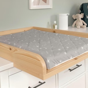 Puede incluir: Una mesa cambiador de madera clara con un cambiador gris con estampado de estrellas. La mesa cambiador está colocada encima de una cómoda blanca. Un bolso para pañales, un peluche y otros artículos para bebés son visibles en el fondo.