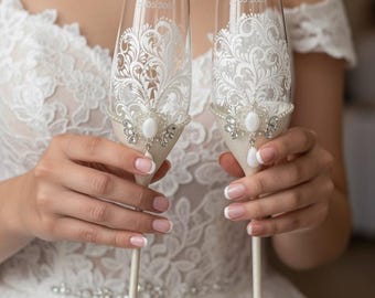May include: Two clear glass champagne flutes featuring white intricate filigree patterns, embellished with a central white oval stone, surrounding clear crystals, and a teardrop accent. The stems are off-white with crystal details at the base.