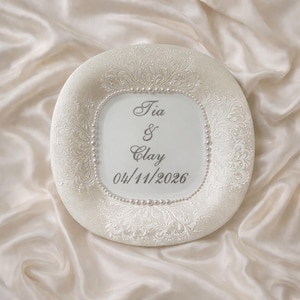 May include: A round white decorative plate featuring an intricate embossed floral pattern on the border, accented with a ring of small white pearls. The center displays the text Tia & Clay and the date 04/11/2026 in a dark grey script font.