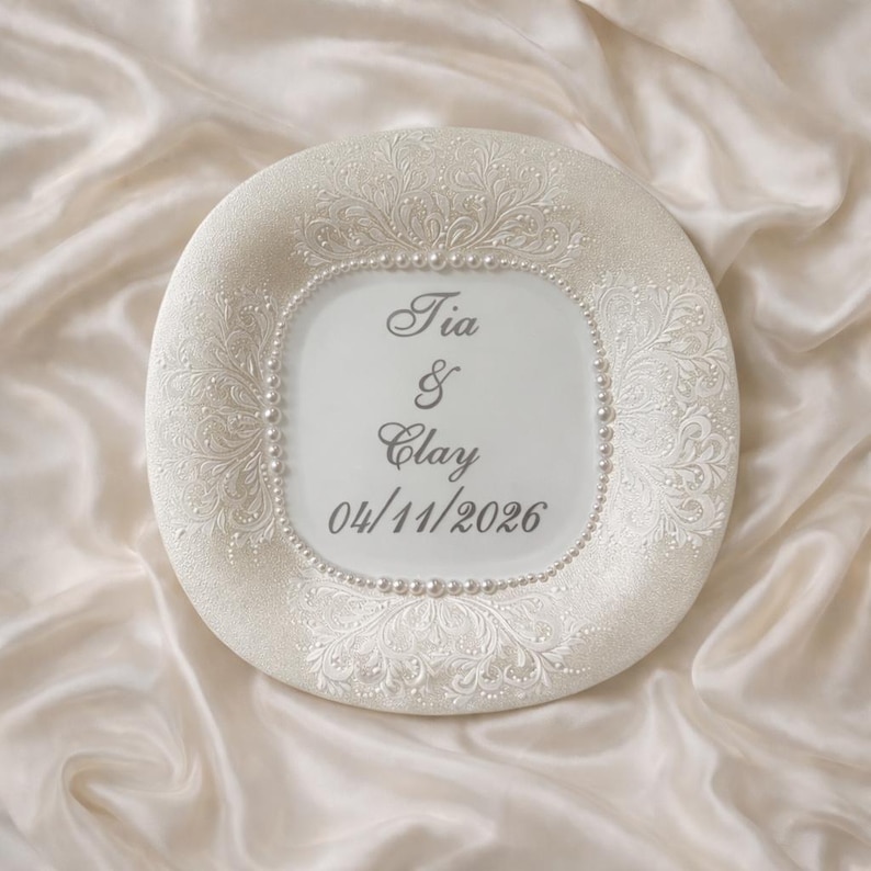 May include: A round white decorative plate featuring an intricate embossed floral pattern on the border, accented with a ring of small white pearls. The center displays the text Tia & Clay and the date 04/11/2026 in a dark grey script font.