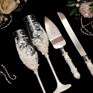 Peut inclure: Ensemble de fl&ucirc;tes &agrave; champagne et de service &agrave; g&acirc;teau pour mariage. Les fl&ucirc;tes sont orn&eacute;es de motifs floraux blancs et de perles. Le serveur et le couteau &agrave; g&acirc;teau ont des poign&eacute;es d&eacute;cor&eacute;es de perles. Le set comprend les noms et la date.