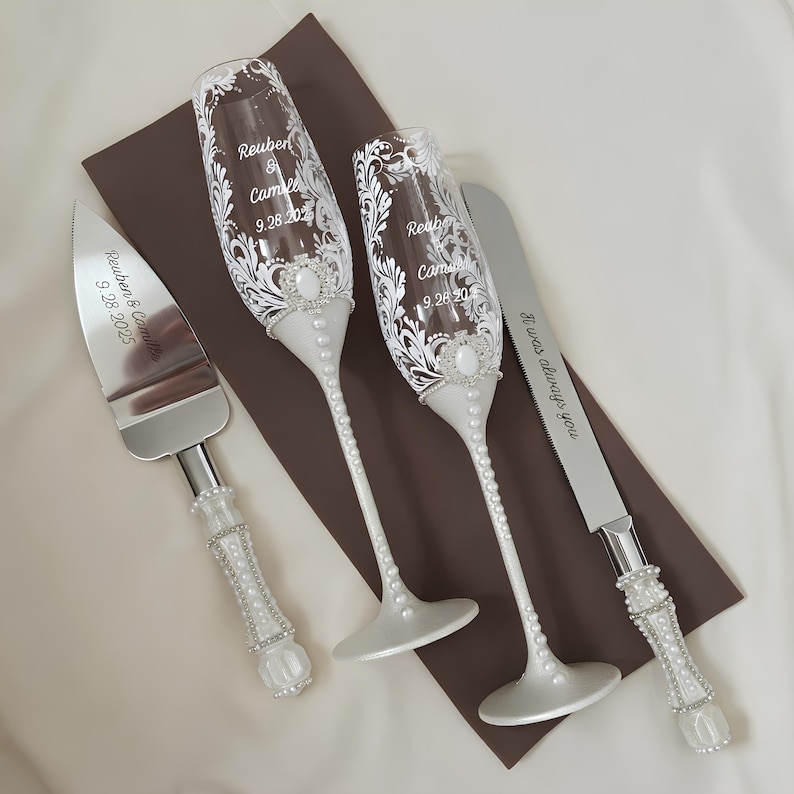 May include: A wedding set featuring two champagne flutes and two serving utensils. The flutes and utensils have white pearl accents and are engraved with names and a date. The text on the knife reads "It was always you."