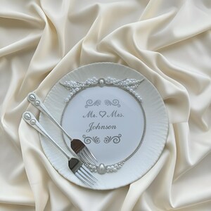 May include: A white plate with a pearl and silver design, featuring the text "Mr. & Mrs. Johnson." Two silver forks are placed on the plate. The plate is set on a cream-colored satin fabric, creating an elegant wedding theme.