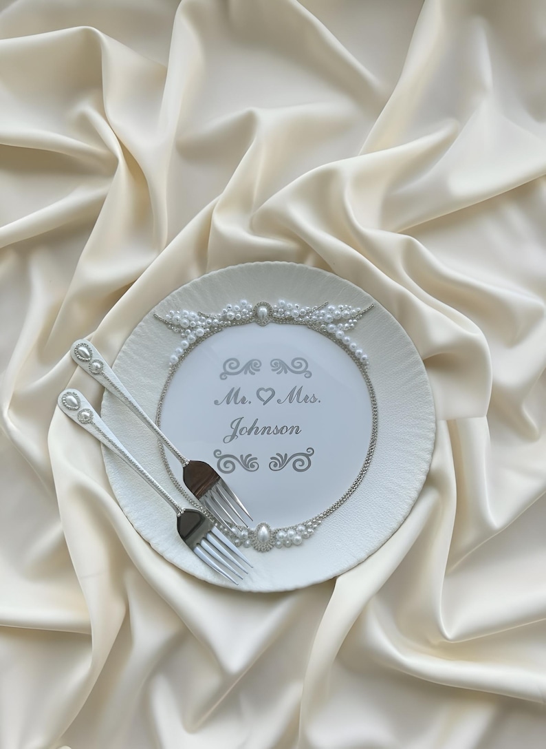 May include: A white plate with a pearl and silver design, featuring the text "Mr. & Mrs. Johnson." Two silver forks are placed on the plate. The plate is set on a cream-colored satin fabric, creating an elegant wedding theme.