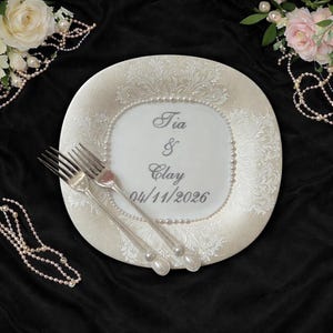 Peut inclure: Une assiette d&eacute;corative blanche avec des motifs floraux et une bordure de perles. L'assiette porte les noms "Tia & Clay" et la date "04/11/2026". Deux fourchettes argent&eacute;es reposent sur l'assiette, avec des accents floraux et perles. Le fond est noir.