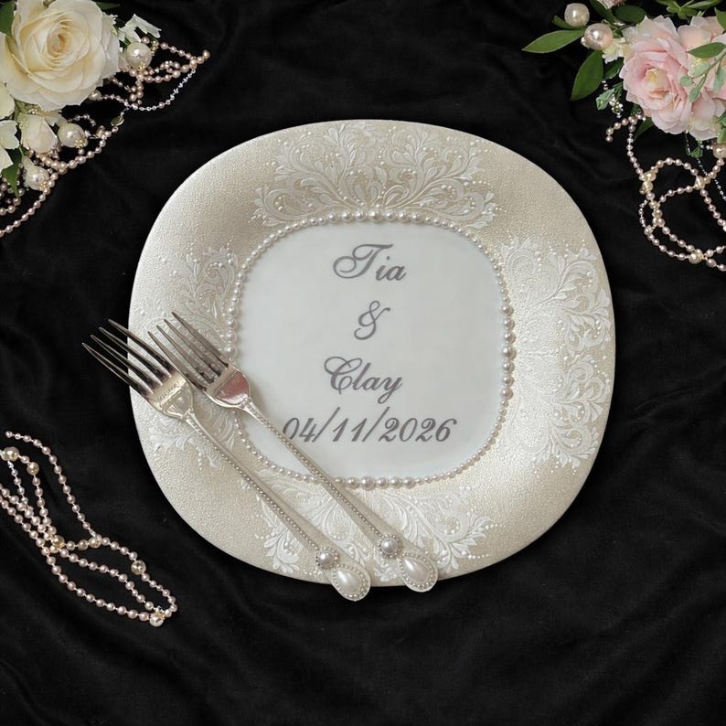 Peut inclure: Une assiette d&eacute;corative blanche avec des motifs floraux et une bordure de perles. L'assiette porte les noms "Tia & Clay" et la date "04/11/2026". Deux fourchettes argent&eacute;es reposent sur l'assiette, avec des accents floraux et perles. Le fond est noir.