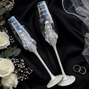 May include: Two clear glass champagne flutes featuring white pearl and silver rhinestone embellishments on the stems and bowls. The glass is etched with the names Isabella and Jackson, a heart symbol, and the date 12. 06. 25 in white script.