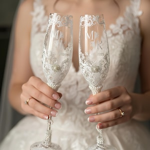 May include: Two champagne flutes with white floral patterns, initials, and the date 08/08/2025. The stems are decorated with pearls and a pearl accent. The flutes are held by a person wearing a wedding dress.