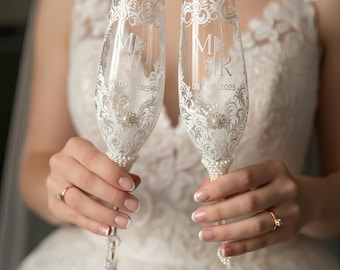 May include: Two clear glass champagne flutes featuring white etched floral scrollwork, the letters M and R, and the date 08.07.2025. Each flute is decorated with a central silver-toned rhinestone flower accent and white pearl beads along the stem.