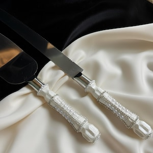 May include: A cake knife and server set with white handles adorned with faux pearls and rhinestones. The stainless steel blades are serrated. The set is displayed on a white and black satin background.