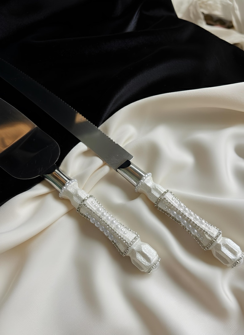 May include: A cake knife and server set with white handles adorned with faux pearls and rhinestones. The stainless steel blades are serrated. The set is displayed on a white and black satin background.
