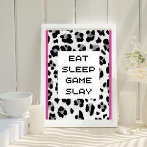 Puede incluir: Una impresión enmarcada en blanco con un fondo de estampado de leopardo blanco y negro. La impresión presenta el texto "EAT SLEEP GAME SLAY" en una fuente pixelada.