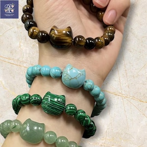 May include: A hand displays four beaded bracelets. The bracelets feature cat-shaped beads in tiger's eye, turquoise, malachite, and jade. The beads are strung with round beads in matching colors. The bracelets are a mix of earth tones and bright colors.