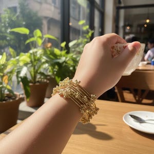 May include: A set of gold-colored bracelets with a beaded design. The bracelets are stacked on a wrist, with a cup of tea in the background. The bracelets have a delicate, ornate appearance.
