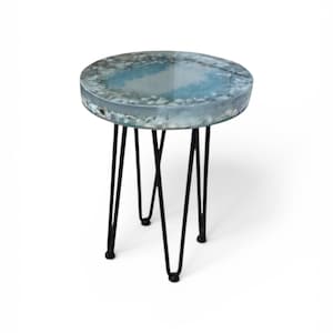 Pode incluir: Uma pequena mesa lateral redonda com um design único. O tampo apresenta um padrão azul e branco, assemelhando-se à água, envolto em resina transparente. Assenta em três pernas pretas de hairpin, criando um visual moderno e elegante.