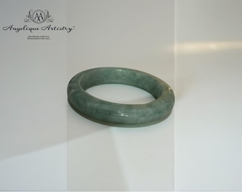 May include: A jade green bangle bracelet. The smooth, oval-shaped bracelet is a muted green colour. The bracelet is displayed on a white surface. The brand name "Angelique Artistry" is in the upper left corner.