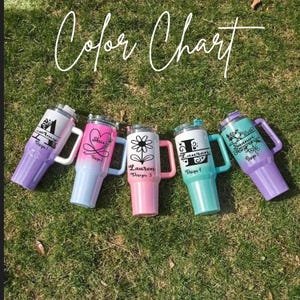 May include: Five insulated tumblers with handles in various colours are arranged on grass. Each tumbler has a unique design and colour scheme, including purple, pink, and teal. The tumblers feature names and floral or graphic designs.