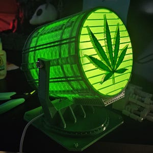 May include: A green, cylindrical lamp with a cannabis leaf design on the front. The lamp is supported by a green stand and base. The lamp is lit with a bright green light, and the cannabis leaf is clearly visible.