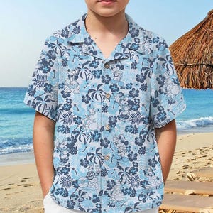 May include: A young person wearing a blue and white patterned short-sleeved shirt with a straw hat. The shirt has a floral design with cartoon characters. The person is standing on a beach.