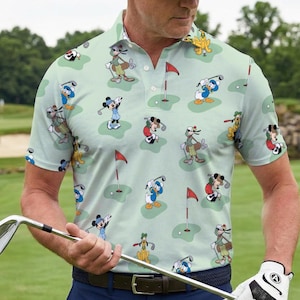May include: Pale green golf polo shirt with a pattern of cartoon characters playing golf. The shirt features Mickey Mouse, Donald Duck, and other characters on a golf course. It has short sleeves and a collared neckline.