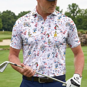 Puede incluir: Polo blanco con un estampado patriótico con personajes de dibujos animados, banderas y fuegos artificiales. La camisa tiene cuello abotonado y mangas cortas. La persona lleva una gorra negra, pantalones azules, un cinturón y un guante de golf, sosteniendo un palo de golf.