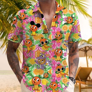 May include: A short-sleeved button-down shirt with a vibrant tropical pattern. The design features cartoon orange characters, flowers, and citrus fruit on a colorful background. The shirt is open, revealing a silver chain.